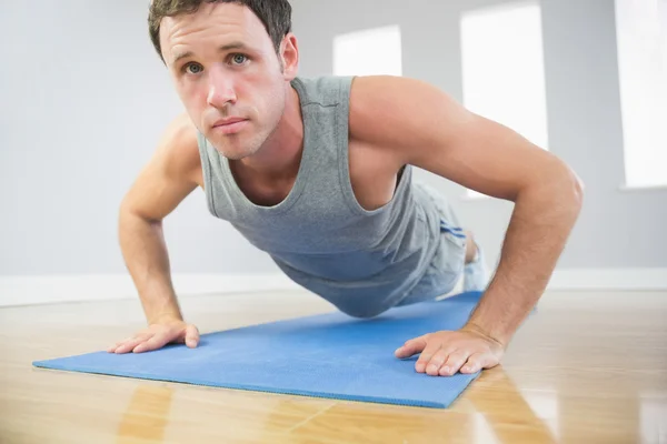 Attractive sporty man doing push ups on blue mat looking at camera — Stock Photo, Image