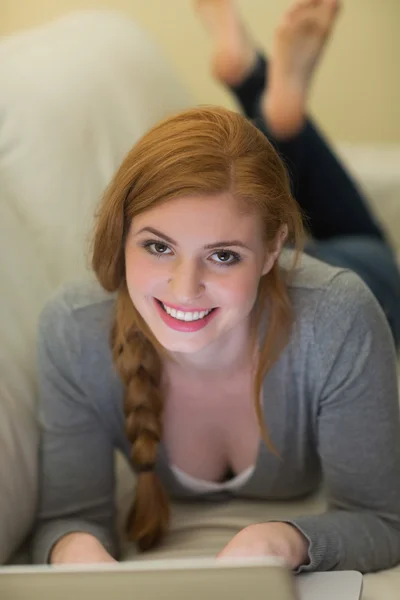 Happy redhead lying on the couch using her laptop at night — Stock Photo, Image