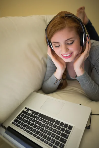 Happy redhead berbaring di sofa dengan laptop mendengarkan musik — Stok Foto