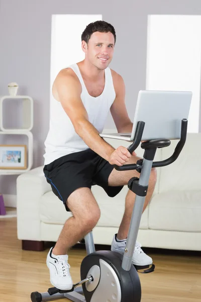 Smiling sporty man exercising on bike and holding laptop — Stock Photo, Image