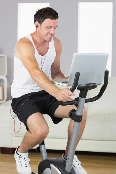 Homem desportivo com fones de ouvido exercitando-se na bicicleta olhando para laptop — Fotografia de Stock