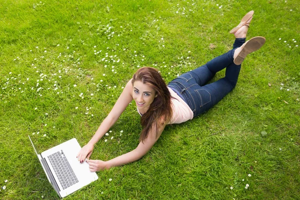 Mujer joven feliz acostada en un césped usando su computadora portátil —  Fotos de Stock