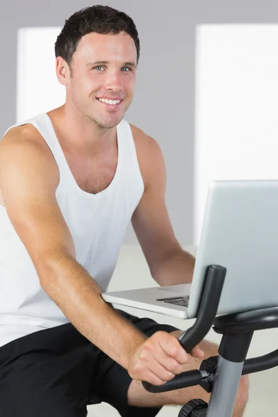 Atractivo hombre deportivo haciendo ejercicio en bicicleta y sosteniendo el ordenador portátil — Foto de Stock