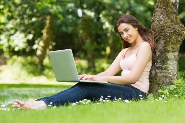 Lachende vrouw leunend tegen een boom in een park met behulp van haar laptop — Stockfoto