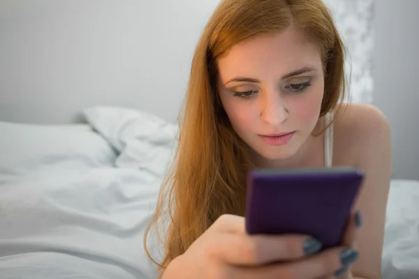 Glad rödhårig liggande på sängen skicka en text — Stockfoto