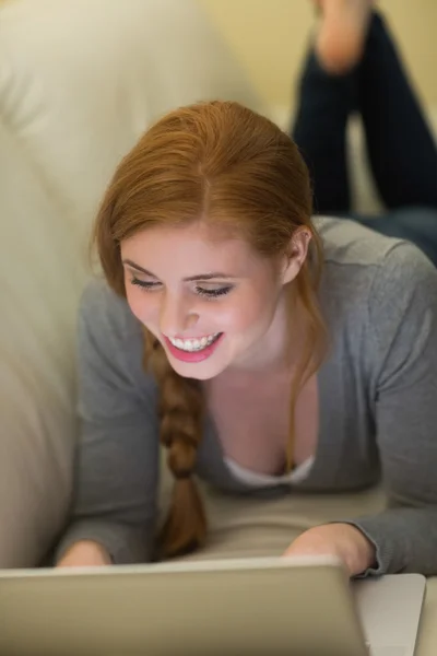 Sonriente pelirroja acostada en el sofá usando su portátil por la noche —  Fotos de Stock