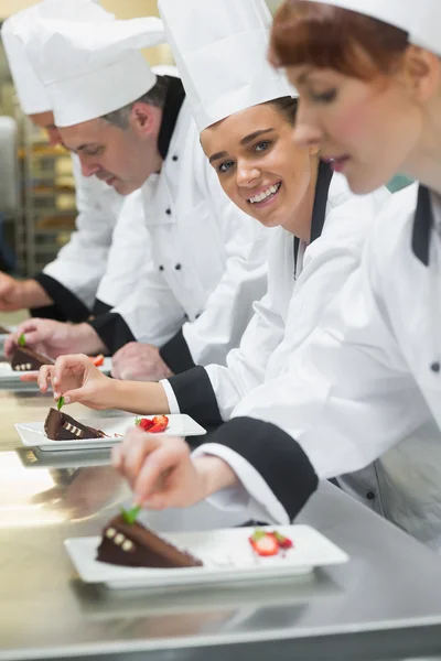 Equipo de chefs en una fila adornando platos de postre una chica sonriendo a la cámara — Foto de Stock