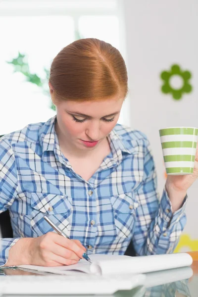 Ruiva escrevendo no bloco de notas em sua mesa e segurando café — Fotografia de Stock