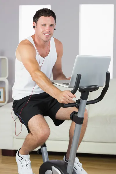 Homem de conteúdo desportivo com fones de ouvido exercitando-se em bicicleta e segurando laptop — Fotografia de Stock