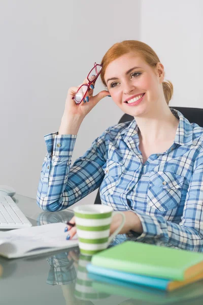 Thoughtful redhead sitting at her desk looking away holding reading glasses — Stock Photo, Image