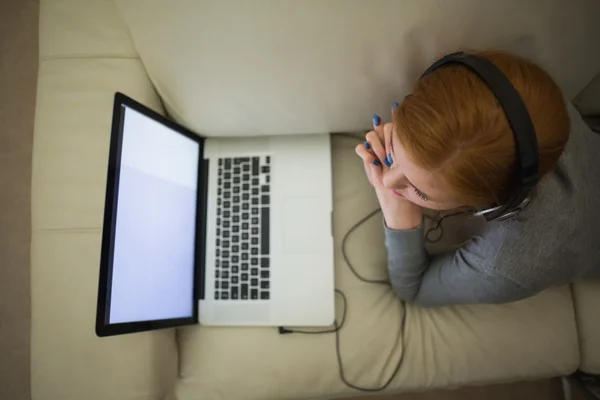 Redhead lying on the couch using her laptop wearing headphones — Stock Photo, Image