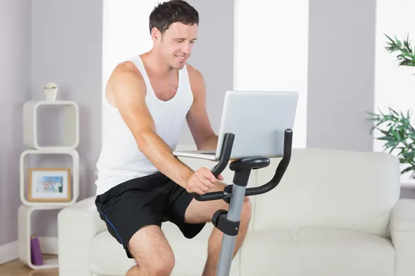 Content sporty man exercising on bike and using laptop — Stock Photo, Image