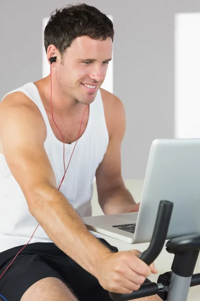Sporty attractive man with earphones exercising on bike looking at laptop — Stock Photo, Image