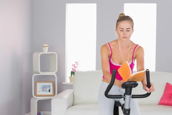 Sportieve gerichte blonde opleiding op hometrainer lezen van een boek — Stockfoto