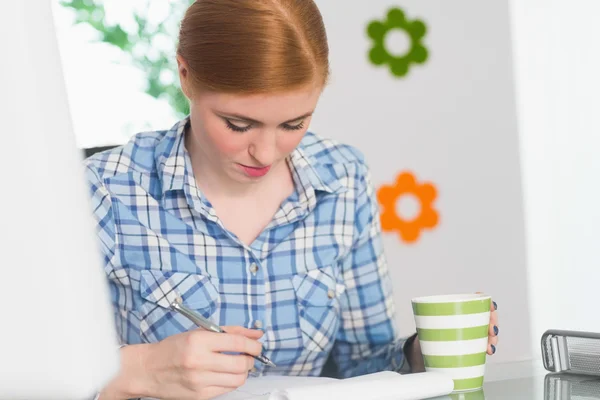 Enfocada pelirroja escribiendo en el bloc de notas en su escritorio y sosteniendo el café — Foto de Stock