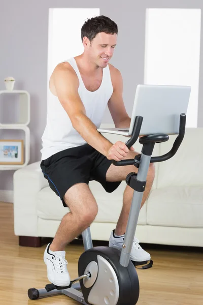 Hombre deportivo feliz haciendo ejercicio en bicicleta y utilizando el ordenador portátil — Foto de Stock