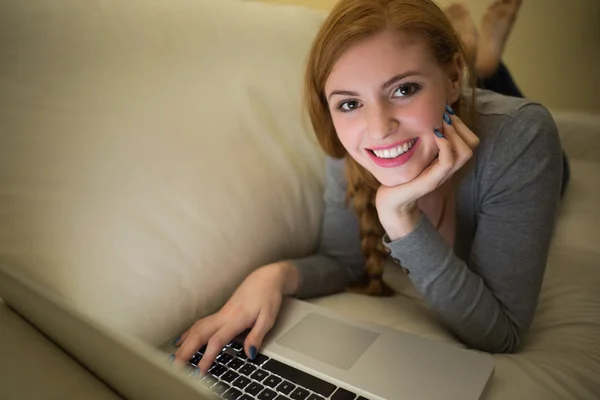 Ruiva sorridente deitada no sofá com seu laptop à noite — Fotografia de Stock