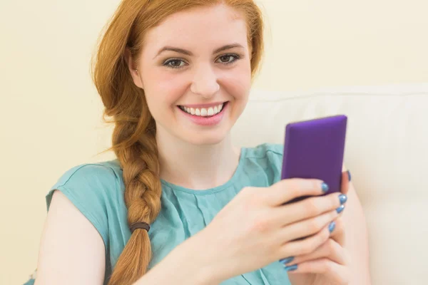 Smiling redhead sitting on the couch using smartphone — Stock Photo, Image