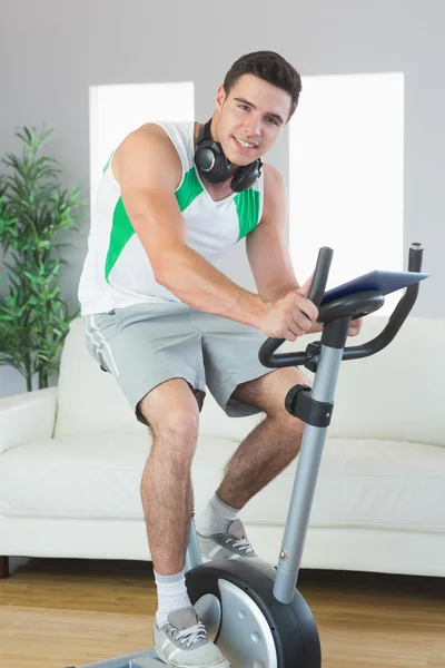 Zufriedener schöner Mann trainiert auf Heimtrainer mit Tablet — Stockfoto