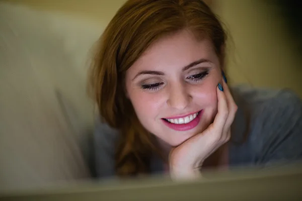 Rossa sorridente sdraiata sul divano guardando il computer portatile — Foto Stock