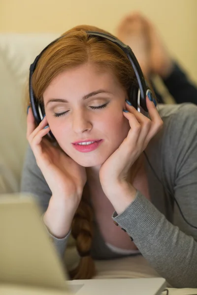 Gadis berambut merah yang tenang terbaring di sofa dengan laptopnya mendengarkan musik. — Stok Foto