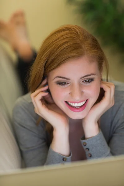 Happy redhead lying on the sofa using her laptop at night — Stock Photo, Image