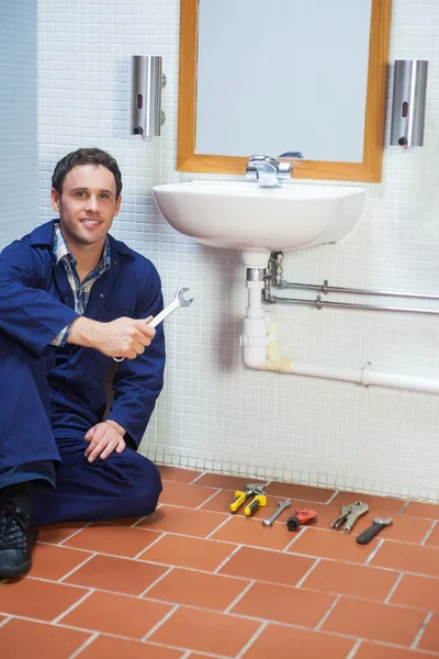 Handsome cheerful plumber sitting next to sink holding wrench — Stock Photo, Image