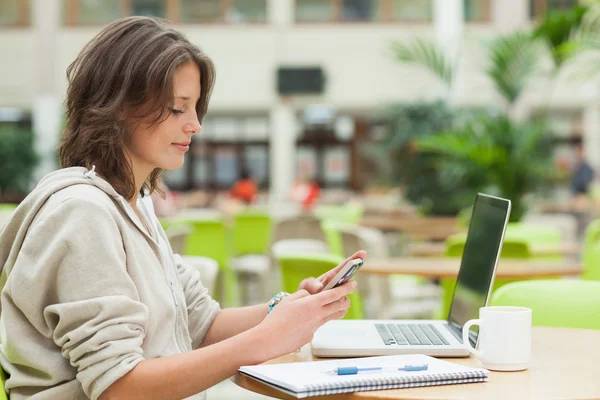 Student using cellphone and laptop at cafeteria table — Stock Photo, Image