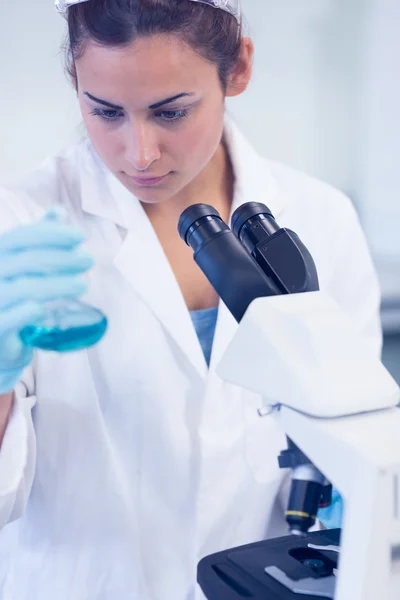 Female researcher examining blue liquid in flask by microscope i — Stock Photo, Image