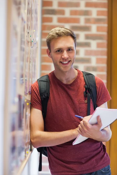 Handsome smiling student taking notes next to notice board — Stock Photo, Image