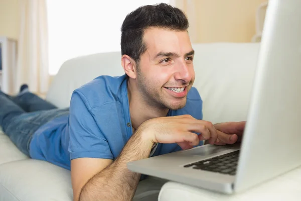 Casual cheerful man sitting on couch using and looking at laptop — Stock Photo, Image