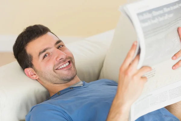 Příležitostné usmívající se muž leží na gauči, drží noviny — Stock fotografie