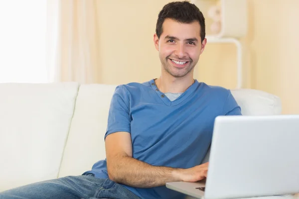 Случайно улыбающийся мужчина сидит на диване с ноутбуком — стоковое фото