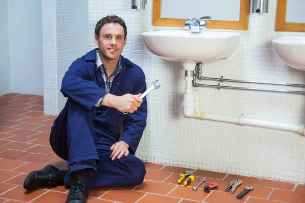 Guapo sonriente fontanero sentado junto al fregadero sosteniendo la llave inglesa — Foto de Stock