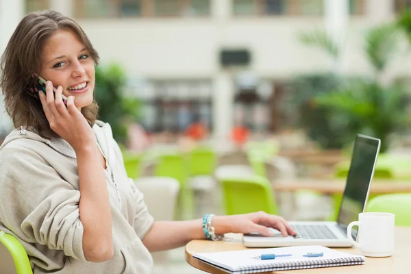 Student using cellphone and laptop at cafeteria table — Stock Photo, Image