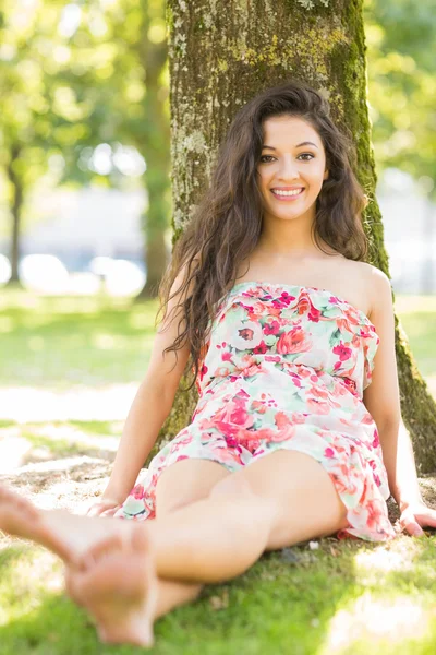 Elegante morena sonriente sentada y apoyada en el árbol —  Fotos de Stock