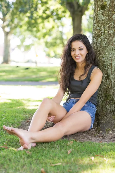 Příležitostné usměvavá brunetka sedí opřený strom — Stock fotografie
