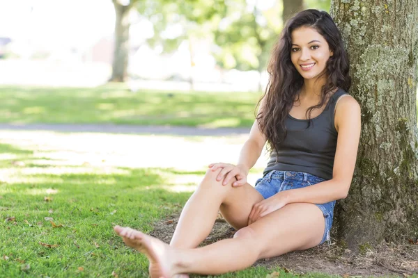Casual attractive brunette sitting leaning against tree — Stock Photo, Image