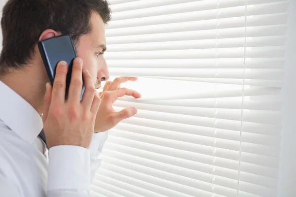 Serious businessman spying through roller blind while phoning — Stock Photo, Image