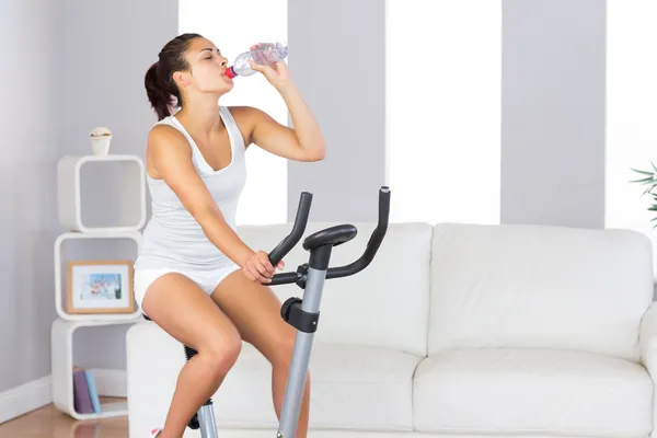 Linda mulher esbelta bebendo enquanto treina em uma bicicleta de exercício — Fotografia de Stock