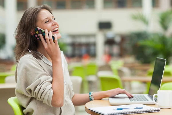 Étudiant joyeux utilisant un téléphone portable et un ordinateur portable à la table de la cafétéria — Photo