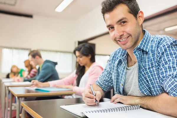 Smiling male student with others writing notes in classroom — Stock Photo, Image