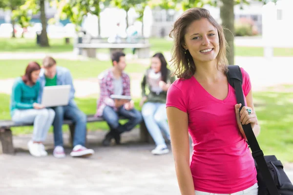 Menina da faculdade sorrindo com alunos desfocados no parque — Fotografia de Stock