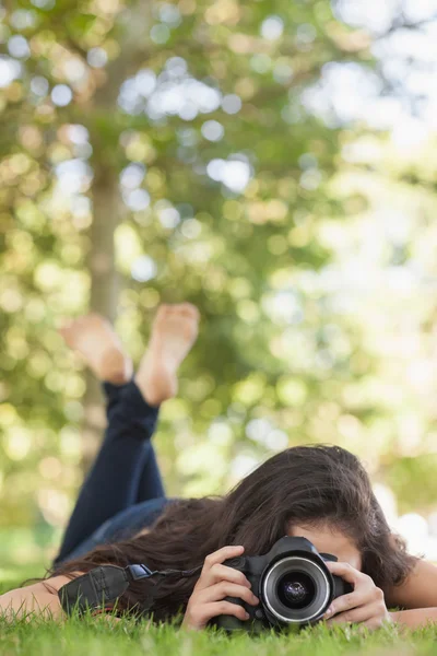 Vista frontal da jovem deitada em um gramado tirando uma foto — Fotografia de Stock