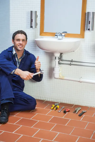 Guapo fontanero alegre sentado al lado del fregadero mostrando el pulgar hacia arriba — Foto de Stock