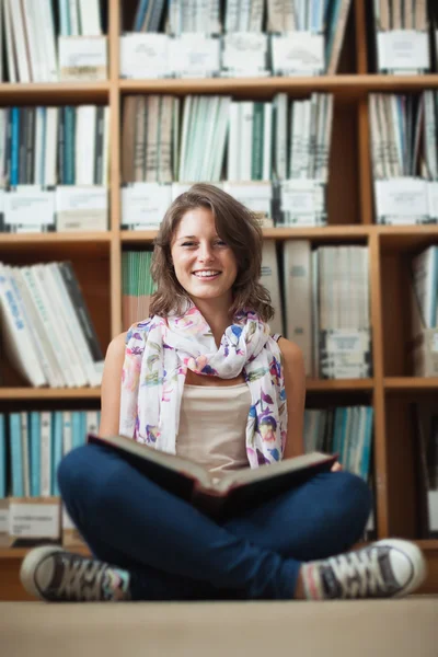 Lachende vrouwelijke student tegen boekenplank lezen van een boek — Stockfoto