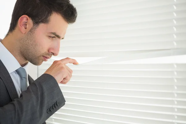 Handsome unsmiling businessman spying through roller blind — Stock Photo, Image