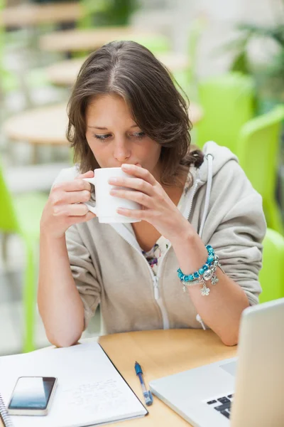 Student drinking coffee while using laptop at cafeteria table — Stock Photo, Image