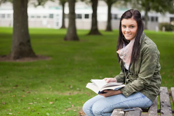 Fröhliche brünette Studentin sitzt auf Bank lesen — Stockfoto