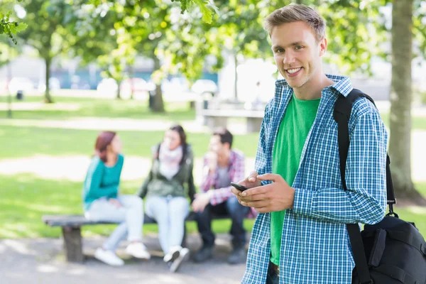 Menino da faculdade mensagens de texto com alunos desfocados no parque — Fotografia de Stock